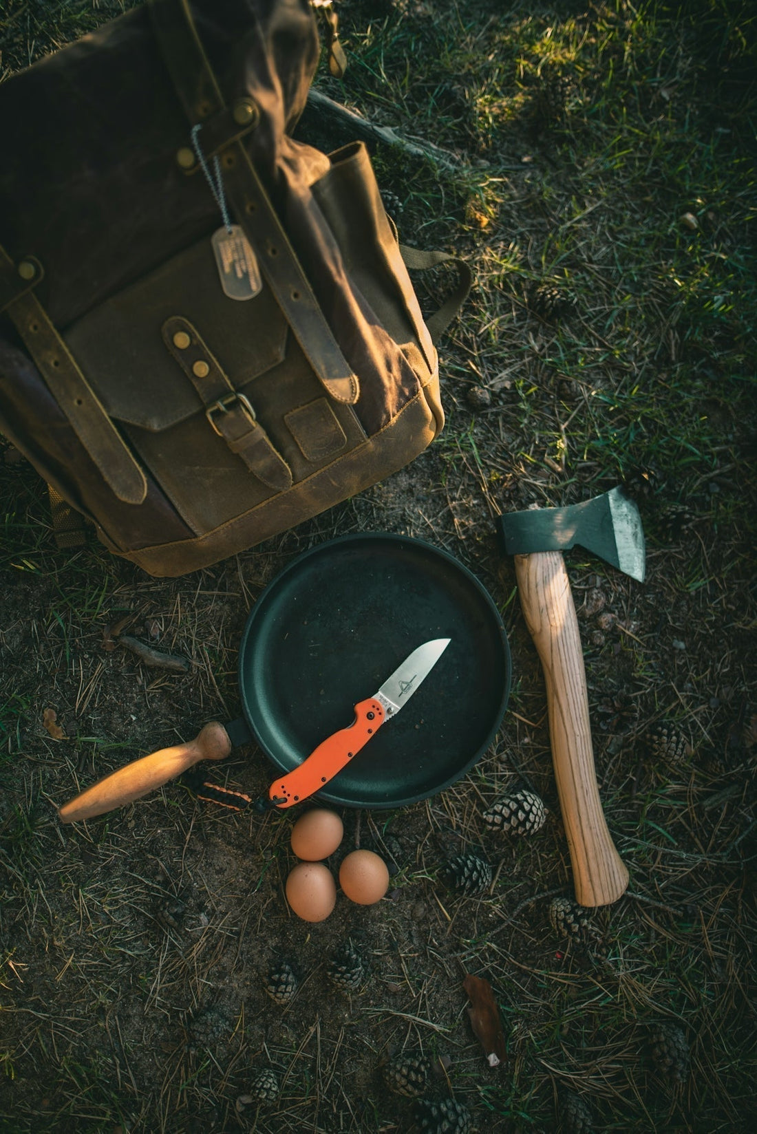 Equipamiento de bushcraft y acampada clásica sobre el suelo del bosque, que incluye una mochila de lona vintage, una sartén de campo con una navaja de mango naranja, un hacha pequeña y tres huevos frescos.