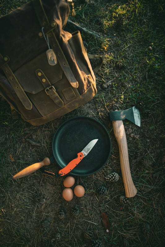 Equipamiento de bushcraft y acampada clásica sobre el suelo del bosque, que incluye una mochila de lona vintage, una sartén de campo con una navaja de mango naranja, un hacha pequeña y tres huevos frescos.
