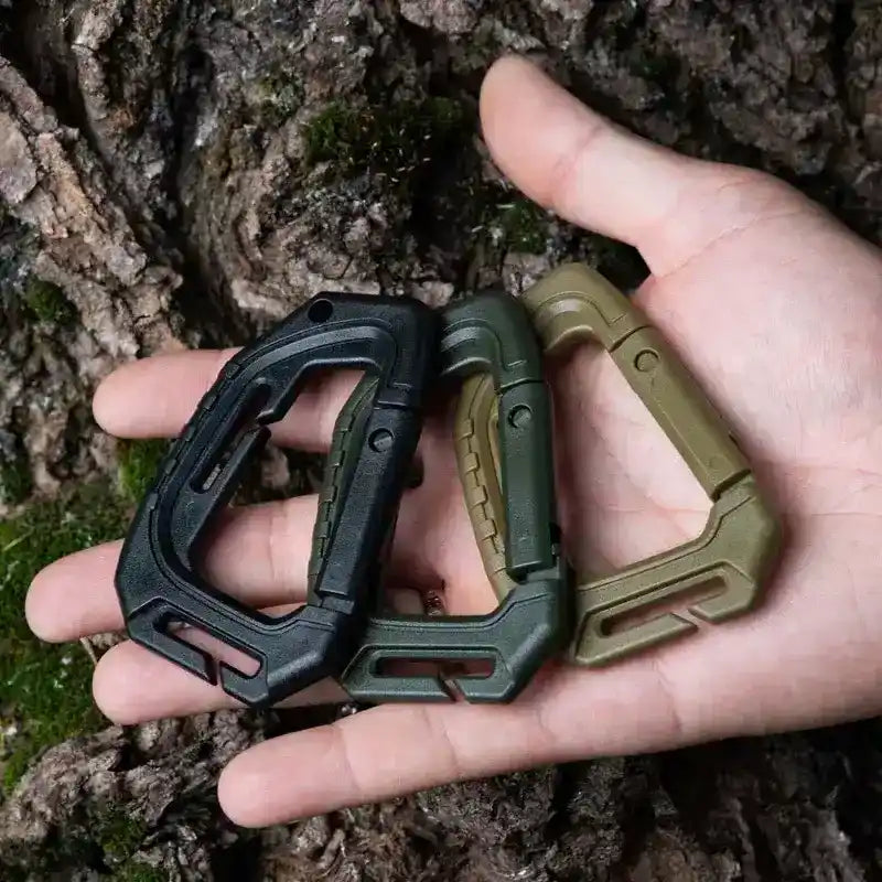 Mano sosteniendo los tres colores del mosquetón táctico Mil-Tec: negro, verde oliva y caqui coyote.